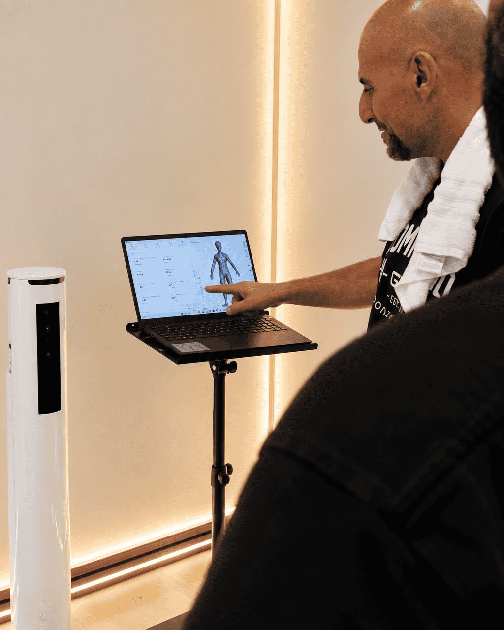 Trainer pointing at a 3D body model on a laptop beside the Styku scanner tower at The Compound Gym