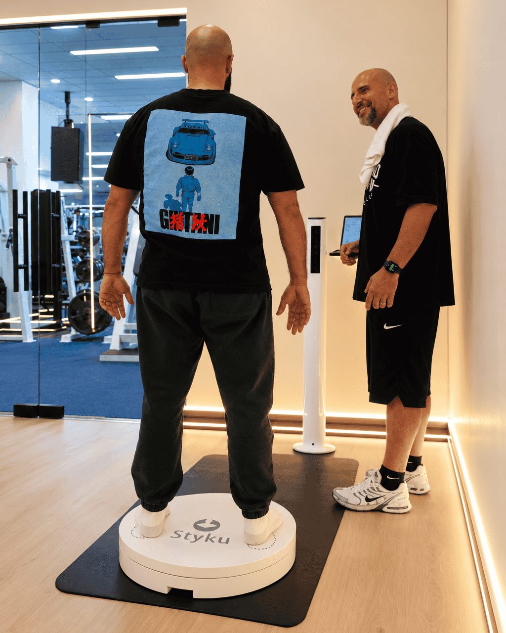 A trainer with a towel over his shoulder smiles beside a client standing on the Styku scanner platform at The Compound Gym