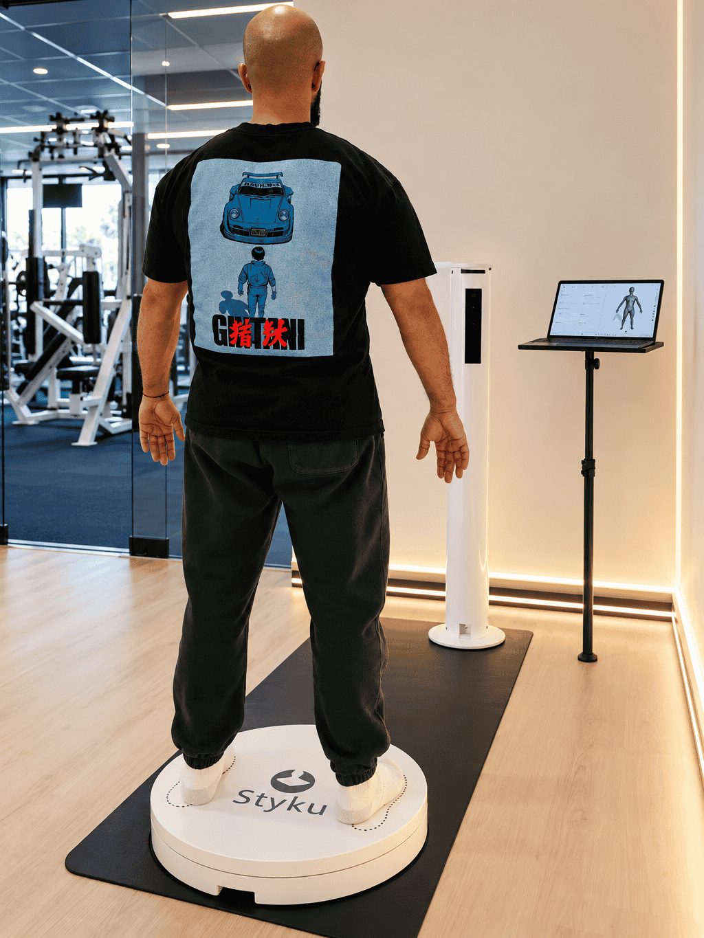 Person standing on the Styku scanner platform in front of the camera tower, with a laptop on a stand showing a 3D body model, at The Compound Gym in Woodland Hills
